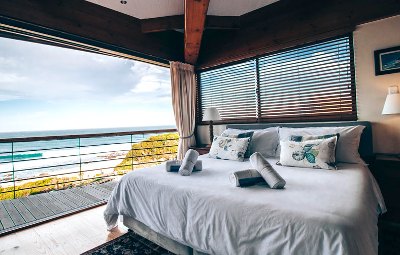 Airbnb room with white bedspread, woven pillows, mountains and a scenic beach view through the window.