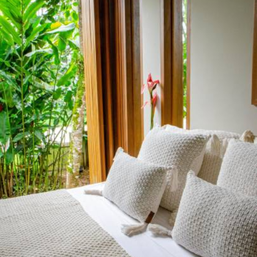 Airbnb bedroom with white bedsheet, woven pillows, beige throw blanket, and large plants on the balcony.