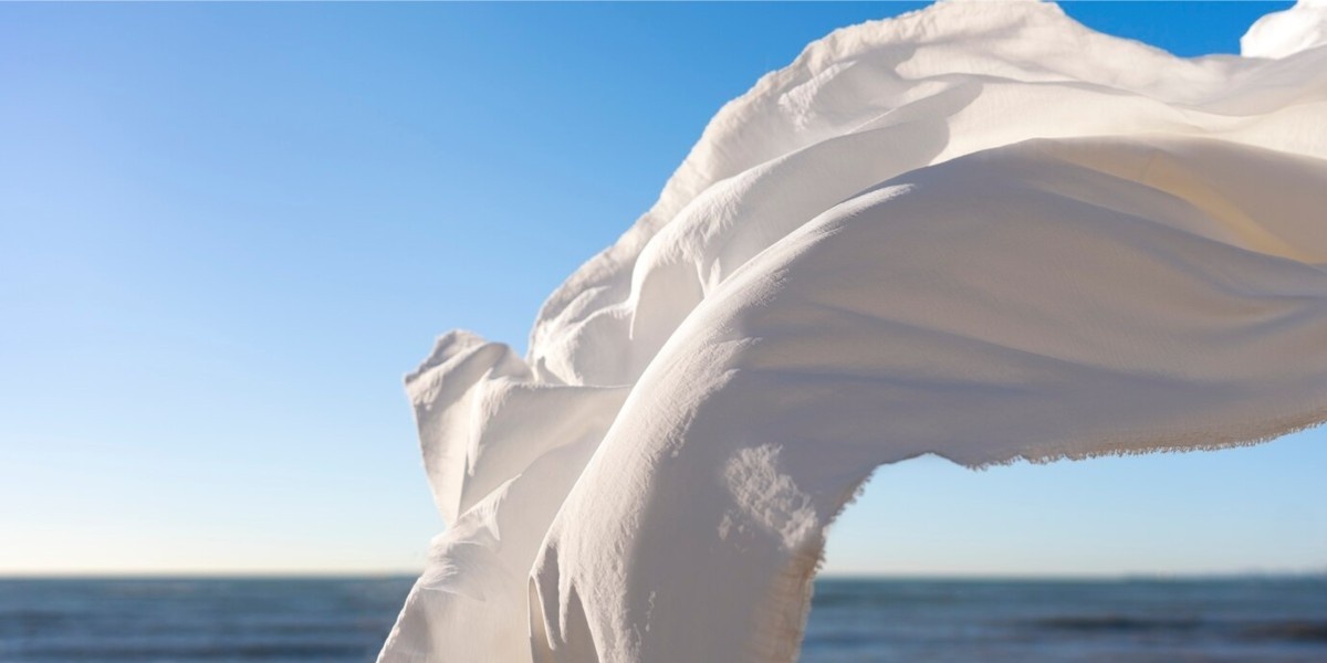 Off-white fabric fluttering in the wind on a sunny beach, with golden sand and clear blue sky in the background.